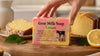A hand holds a bar of Goat Milk Soap Lemon in a pink and yellow package, set on a wooden cutting board with sliced lemonade cake, a lemon half, and green leaves in a bright kitchen setting.