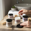 Person applying cream from a jar on a bathroom counter with various skincare products.