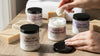 Person applying cream from a jar on a bathroom counter with various skincare products.
